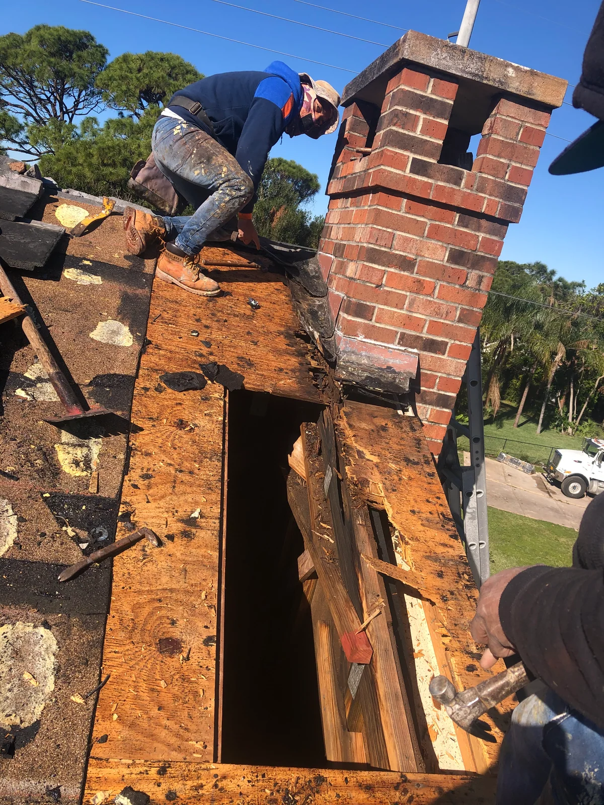 Opened tile roof repair area beside a brick chimney, with deteriorated wood decking and a section removed.